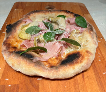 Homemade Neapolitan-style pizza topped with thinly sliced mortadella, marinated artichokes, and fresh basil leaves on a golden, charred crust.