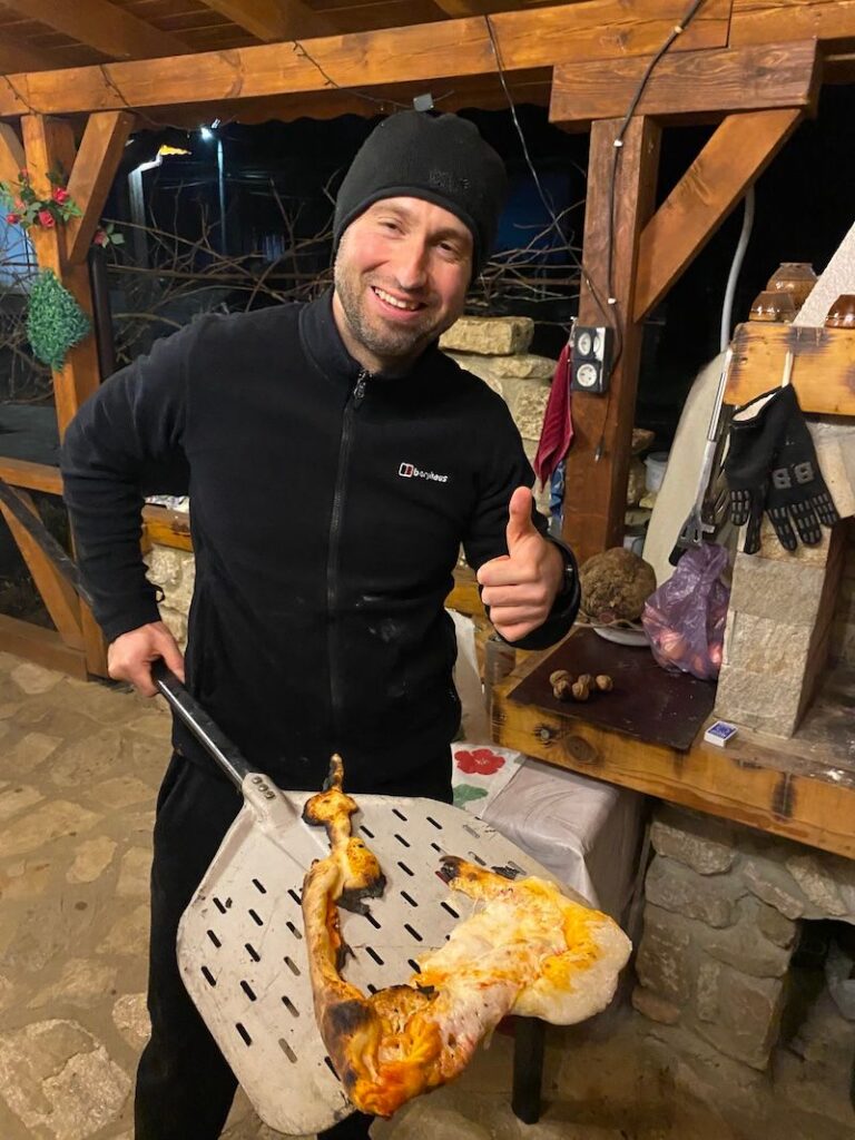 A disappointed yet determined home cook holds up a misshapen, partially fallen-apart pizza fresh from a wood-fired oven.