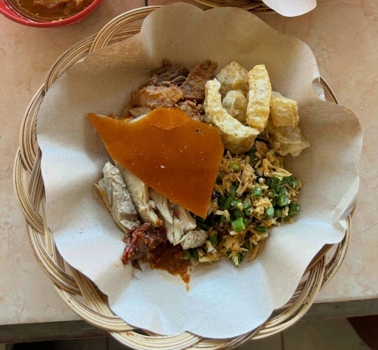 A plate of Babi Guling in Padangbai, Bali, featuring crispy golden pork skin, tender spiced meat, lawar (vegetable mix), soto (pork soup), and sambal on the side."