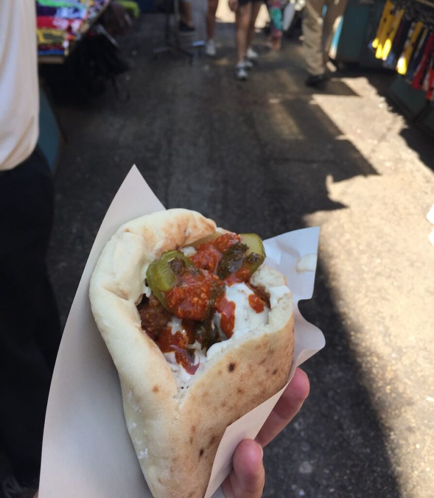 A freshly made falafel pita sandwich filled with crispy falafel balls, vibrant chopped salad, and drizzled with tahini, held in hand against the lively backdrop of Carmel Market in Tel Aviv.
