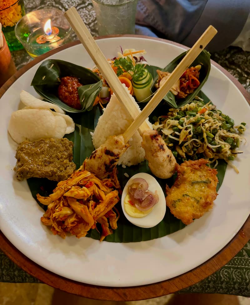 A beautifully presented plate of Nasi Campur in Ubud, Bali, featuring steamed rice surrounded by a variety of side dishes, including grilled chicken, crispy tempeh, sautéed vegetables, sambal, and a flavorful Balinese curry.