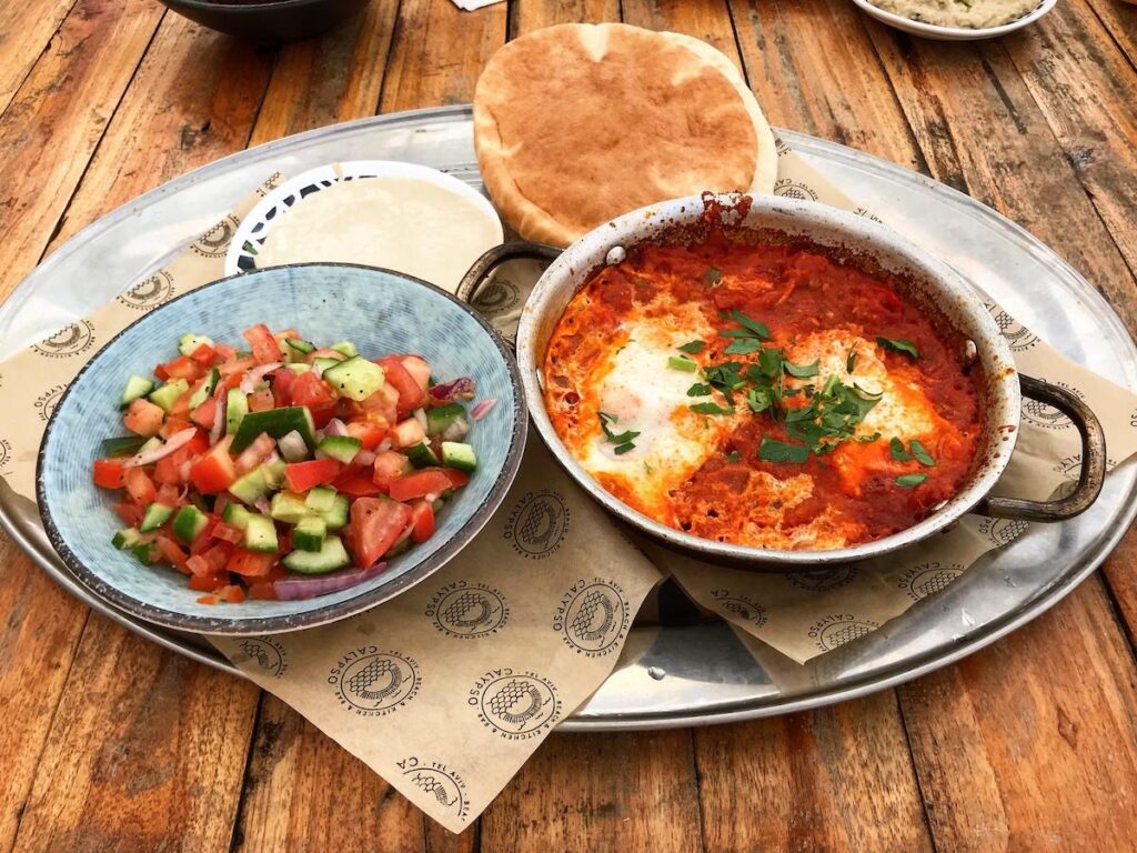 A vibrant pan of shakshuka with poached eggs in a spiced tomato and bell pepper sauce, garnished with fresh herbs. Served alongside warm, fluffy pita bread and a crisp salad with cucumbers and tomatoes.