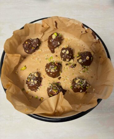 Homemade raw cherry pistachio hazelnut balls coated in coconut shavings and topped with dark chocolate ganache, garnished with crushed nuts, served on a tray.