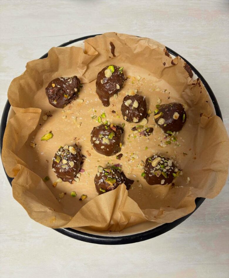 Homemade raw cherry pistachio hazelnut balls coated in coconut shavings and topped with dark chocolate ganache, garnished with crushed nuts, served on a tray.